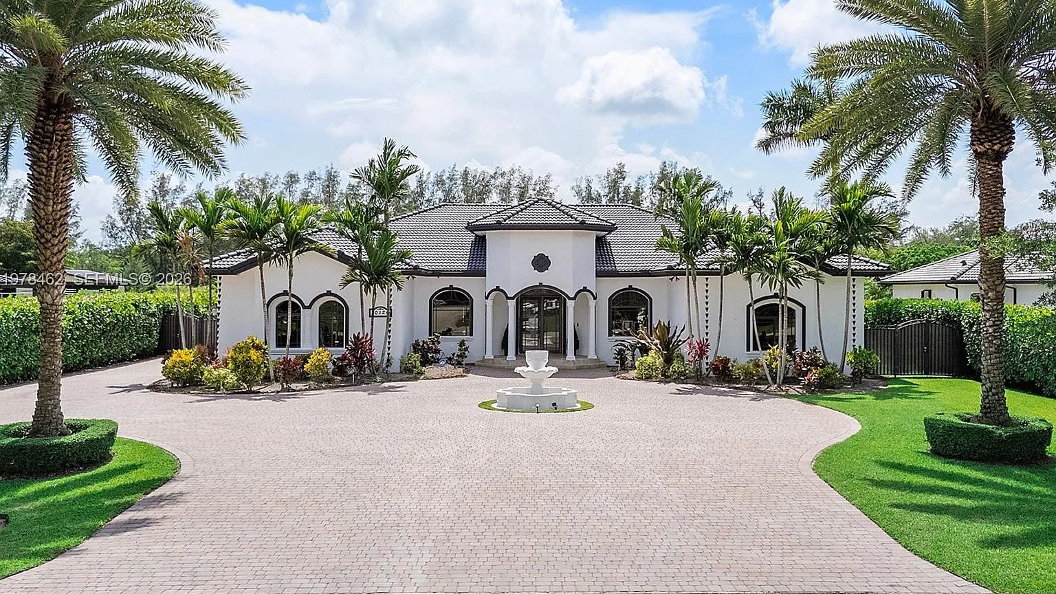 Real MLS listing photo: white Mediterranean estate with a circular paver driveway, fountain and palm trees